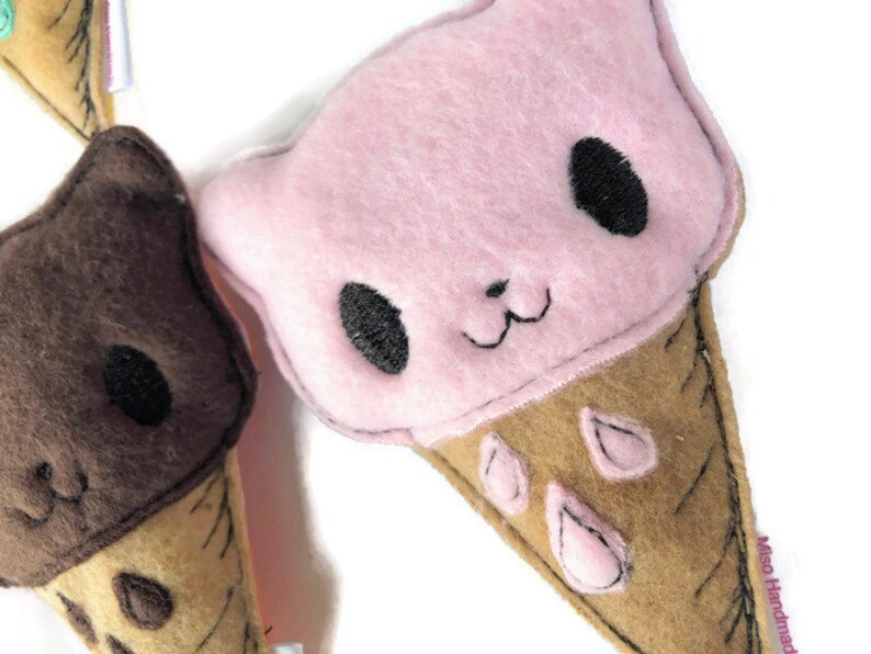 May include: Two felt ice cream cone plushies. One is pink with black eyes and a brown cone. The other is brown with black eyes and a tan cone.