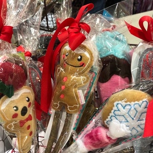 May include: A display of festive, handcrafted ornaments, including gingerbread men, cupcakes, and snowflake designs, all attached to wooden sticks and wrapped in clear cellophane. Each ornament is adorned with red ribbon.