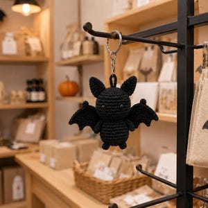 May include: A black crocheted bat keychain featuring a round body, pointed ears, and outstretched wings. The bat is crafted from yarn with a visible stitch pattern and includes two small black bead eyes. It hangs from a silver metal keyring.