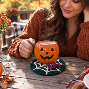 May include: A round orange ceramic mug shaped like a jack-o'-lantern with a black carved face, resting on a dark green circular crochet coaster featuring a white spiderweb pattern and a small purple crocheted spider attached to the surface.