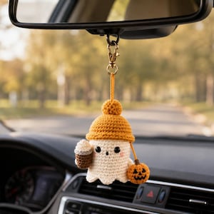 May include: A crocheted ghost car charm hanging from a rearview mirror. The ghost is off-white with black eyes, pink cheeks, and a mustard yellow beanie. It holds a brown coffee cup in one hand and a small orange jack-o'-lantern in the other.