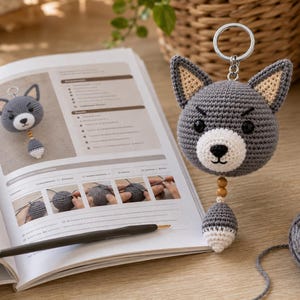Puede incluir: Un llavero de cabeza de lobo tejido a ganchillo en color gris con ojos negros, hocico blanco y orejas puntiagudas con centros beige. Una pequeña cola gris y blanca cuelga debajo de la cabeza, unida por dos cuentas de madera. Un anillo de metal plateado está sujeto en la parte superior.