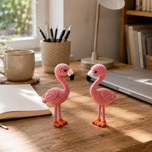 Puede incluir: Dos flamencos de ganchillo hechos a mano con cuerpos de hilo rosa brillante, cuellos largos y patas naranjas. Cada flamenco tiene un detalle de pico blanco y negro. Las figuras están tejidas con un patrón de puntos visible y se mantienen en pie.
