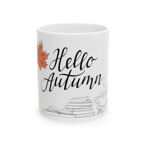 Op de afbeelding: Een witte keramische mok met de zwarte cursieve tekst Hello Autumn. Het ontwerp op de voorzijde bevat een enkel oranje esdoornblad, een stapel boeken en een lijntekening van een dampende beker of kan voor warme dranken.