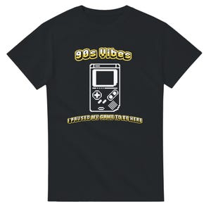 Könnte beinhalten: Ein schwarzes Kurzarm-T-Shirt mit einer weißen, pixeligen Grafik einer tragbaren Spielkonsole. Über der Konsole steht der gelb-weiße Text 90s VIBES und darunter der gelb-weiße Text I PAUSED MY GAME TO BE HERE.
