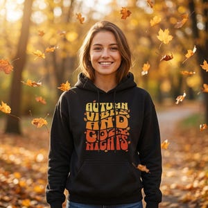 May include: A black hooded sweatshirt featuring a graphic print in yellow, orange, and red. The text reads "AUTUMN VIBES AND COZY NIGHTS" in a stylised, wavy font, accented with small leaf illustrations scattered throughout the lettering.