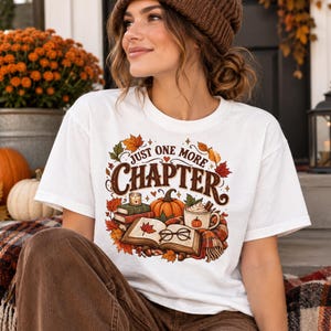 Puede incluir: Una camiseta blanca con un estampado gráfico colorido con el texto "JUST ONE MORE CHAPTER" rodeado de hojas de otoño, una pila de libros, una calabaza, una vela, una taza de chocolate caliente y unas gafas en tonos cálidos de temporada.