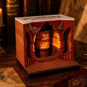 May include: A 3D paper carving art piece featuring a red and orange layered structure with a central illuminated tower. The top white panel displays the text "HOGWARTS". Intricate cutouts include a bird and a hat silhouette within the archway.