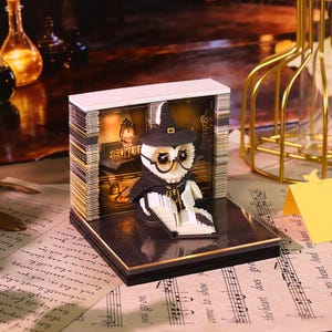 May include: A 3D paper craft diorama featuring a white owl wearing a black witch hat and round glasses, reading an open book. The owl is positioned in front of a backdrop with the text "CENTURY" and a vintage library illustration, all set on a black base.