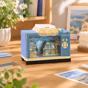 Könnte beinhalten: Ein rechteckiges, blaues Diorama in Form eines Fernsehers mit einer Miniatur-Straßenszene und Gebäuden im Inneren. Der Rahmen besteht aus geschichtetem Papier mit einem weißen Oberteil, einem schwarzen Sockel und einem runden Bedienknopf auf der rechten Seite.