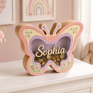 Può includere: Un salvadanaio in legno a forma di farfalla con un pannello frontale trasparente, che riporta il nome Sophia in corsivo. La cornice è dipinta in rosa, giallo e viola pastello con motivi floreali e l'interno è riempito con monete color oro.