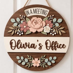 Può includere: Una targa da porta circolare in legno con finitura marrone scuro che riporta il testo "IN A MEETING" e "Olivia's Office" in lettere marroni. La targa è decorata con ritagli floreali a strati nei toni del rosa, bianco e verde salvia.