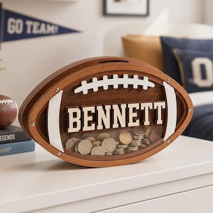 May include: A wooden football-shaped money box with a clear front panel showing a collection of coins inside. The name BENNETT is displayed in white block letters across the centre, with white laces above and white accents on the sides.
