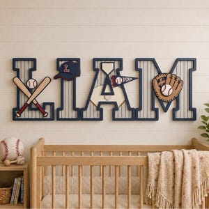 Op de afbeelding: Een houten wandbord met de naam LIAM in grote marineblauwe letters met witte krijtstrepen. De letters zijn voorzien van uitsparingen met een honkbalthema, waaronder een honkbalpet, gekruiste knuppels, een honkplaat, een wimpel en een handschoen.