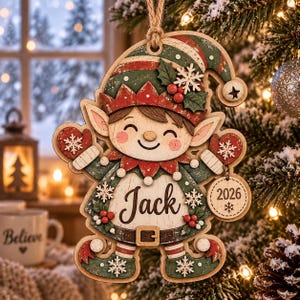May include: A wooden Christmas tree ornament shaped like an elf wearing a green and red hat with a white snowflake and holly berries. The elf has red mittens, a belt, and a round tag that says 2026. The center of the elf features the name Jack.