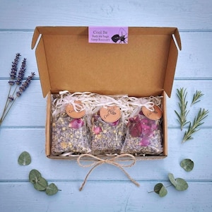 May include: A brown cardboard box containing three clear plastic bags filled with dried flower petals and herbs. Each bag has a round tan tag with a bee illustration and the text Cool Be. A purple label on the box reads Cool Be Bath tea bags Sleep Recovery.
