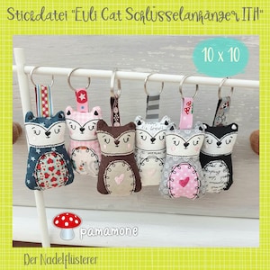 May include: Seven colorful fabric cat keychains hanging from a white metal rack. Each keychain features a different fabric pattern and a cute embroidered cat face. The text "Stickdatei "Euli Cat Schlüsselanhänger ITA" and "10 x 10" is visible in the image.
