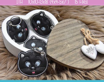 May include: Black cat face keychains made of textured material with embroidered blue eyes, white whiskers, and a pink tongue. The keychains feature silver metal rings and are displayed in a white heart-shaped container with a wooden lid.