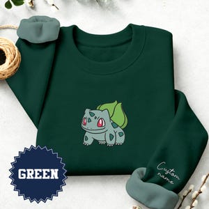 Puede incluir: Una sudadera de cuello redondo verde oscuro con una criatura bordada de color verde claro que tiene un bulbo verde grande en la espalda y ojos rojos. La manga incluye el texto Custom name y una insignia azul oscuro muestra la palabra GREEN.