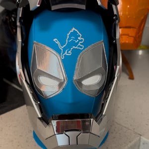 May include: A full-face helmet featuring a bright blue base color with a metallic silver faceplate and trim. The forehead area displays a white outline graphic of a lion in a leaping pose. The design includes angular eye cutouts and a chin guard.
