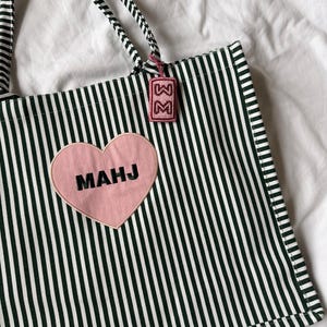 May include: A tote bag featuring a dark green and white vertical striped pattern. A pink heart-shaped patch is centered on the front with the black text MAHJ. A small rectangular pink beaded tag with the letter M hangs from the striped handle.