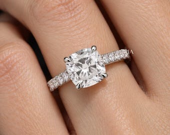 Könnte beinhalten: Ein silberfarbener Ring mit einem großen, kissenförmigen Mittelstein, der von vier Krappen gehalten wird. Die Ringschiene besteht aus Metall und ist mit einer Reihe kleinerer, rund geschliffener Steine besetzt. Das Wort ORIA ist sichtbar.