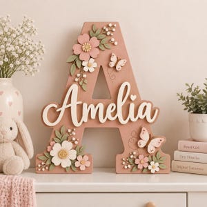 Könnte beinhalten: Ein großer, altrosa Holzbuchstabe A mit dem Namen Amelia in weißer Schreibschrift. Der Buchstabe ist mit geschichteten Papierblumen in Rosa- und Weißtönen, grünen Blättern und zwei Schmetterlingen mit gepunkteten Flugbahnen verziert.