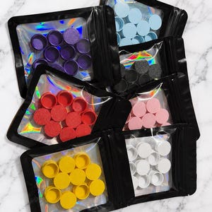 May include: A collection of six black resealable pouches with clear windows, each containing multiple small circular objects in distinct solid colors: light blue, purple, black, red, light pink, yellow, and white, arranged in a cluster.