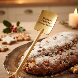 Op de afbeelding: Een goudkleurige metalen lepel in de vorm van een schep met een platte, vierkante kop. De tekst "Michael's Christmas Cookie Spoon" is in de kop van de lepel gegraveerd. Het handvat heeft een driehoekige opening aan het uiteinde voor grip.