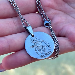 May include: A silver-toned circular pendant necklace featuring a laser-engraved illustration of Saint Rita holding a crucifix. The pendant is attached to a silver-toned rolo chain. The text St. Rita is clearly engraved on the lower right side.