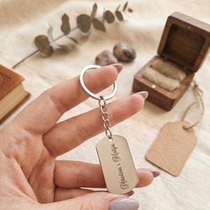 May include: A silver-toned metal dog tag keychain featuring a split ring and a short chain. The rectangular tag has rounded corners and is engraved with the text "Nikolina i Matija" in a cursive script font, positioned vertically on the surface.