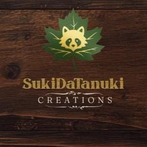 May include: A logo featuring a gold tanuki face centered on a green maple leaf. Below the leaf, the text SukiDaTanuki CREATIONS is written in gold lettering. The entire design is set against a dark brown, horizontal wood grain background.