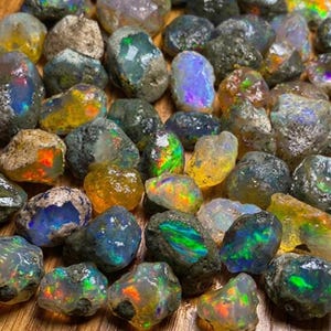 May include: A collection of raw, unpolished opal stones featuring irregular shapes and textures. Each stone displays a unique play of color, including flashes of neon green, vibrant orange, deep blue, and yellow, embedded within dark, earthy rock matrices.