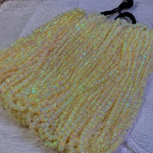 Puede incluir: Una colección de múltiples hilos de pequeñas cuentas redondas de color amarillo pálido con un acabado iridiscente. Las cuentas están dispuestas en filas paralelas y aseguradas en la parte superior con lazos de hilo negro, creando un aspecto uniforme.