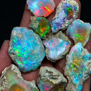 May include: A collection of raw, unpolished opal stones featuring vibrant flashes of iridescent colour including neon green, electric blue, fiery orange, and bright red against a natural, textured, light-coloured rocky matrix base.
