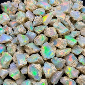 Könnte beinhalten: Eine Sammlung roher, unpolierter Opalsteine mit einer milchig weißen Grundfarbe und lebhaften Blitzen aus irisierendem grünem, orangefarbenem und rosa Licht. Jeder Stein hat eine raue, unregelmäßige Textur mit natürlichen Kanten.