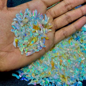 May include: A large collection of small, irregular, translucent white opal chips featuring iridescent flashes of blue, green, yellow, and pink. These polished gemstone fragments exhibit a smooth, glossy texture and are scattered across a dark surface.