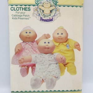May include: A Butterick sewing pattern envelope for Cabbage Patch Kids Preemies, number 3269. The package features three dolls wearing handmade clothes: a pink dress, a yellow shirt and pants set, and a white patterned romper with matching booties.