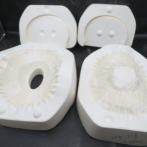 May include: Two white ceramic slip casting molds for pottery. The left mold features a hollow center with a ruffled edge, while the right mold displays a raised, textured floral or ruffled pattern. Both pieces have small circular indentations.