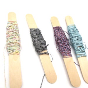 May include: Four wooden craft sticks each wrapped with colorful metallic thread. The thread colors include a rainbow mix, silver, purple and blue. The craft sticks are arranged on a white background.