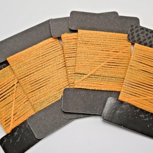 May include: Five rectangular black cardboard bobbins wrapped with shimmering orange metallic thread. The bobbins feature a dark gray animal print pattern on the top and bottom edges. The orange thread is wound horizontally across the center.