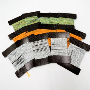 May include: A collection of spools of thread in various colors, including silver, orange, and green. Each spool is attached to a black card with a reptile-like pattern. The threads appear to be for crafting or decorative purposes.