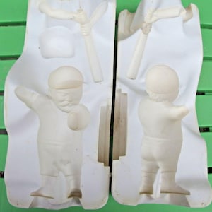 May include: Two-part white ceramic mold featuring a baseball player figurine. The mold includes details for the player's uniform, cap, and a baseball bat. Additional elements include a baseball and a separate cap. The mold is designed for creating ceramic figurines.