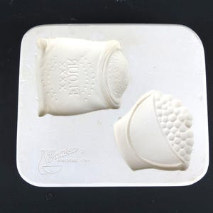 May include: A white ceramic mold featuring two detailed designs: a sack with the word "HOOK" and a bowl of textured, round shapes. The mold is set on a white rectangular base, with the "Duncan Enterprises" logo visible.