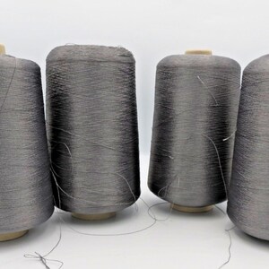 May include: Four large industrial spools of metallic silver-grey thread arranged in a row. Each spool features a cylindrical core with a flat top and bottom, densely wound with fine, shimmering thread that displays a subtle, reflective texture.