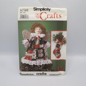 May include: A Simplicity Crafts 9796 sewing pattern envelope featuring a doll with brown hair wearing a patchwork dress with red, black, and white patterns, and a matching Christmas stocking. The text includes Simplicity Crafts and 9796.