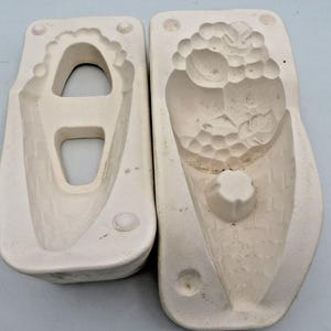 May include: Two off-white ceramic molds. One mold has a large opening and a rounded base. The other mold has a textured surface with a floral design and a small, round protrusion at the base. Both molds are rectangular.