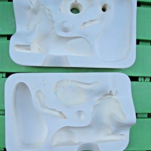 May include: Two white silicone molds, each containing the detailed forms of a reclining unicorn. The molds are rectangular with various cavities for the unicorn's body parts, including the head, legs, and body. The molds are designed for crafting.