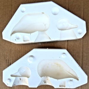 May include: Two white, duck-shaped molds. Each mold has a triangular shape with rounded corners and contains two duck profiles. The molds are made of a solid, opaque material and are likely used for casting or creating duck-shaped objects.