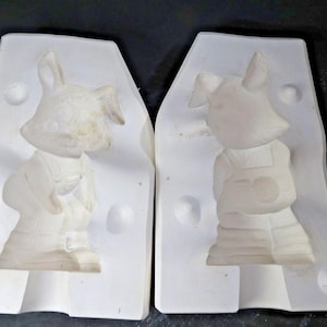 May include: Two white ceramic molds, each depicting a cartoon bunny wearing overalls. The bunny has large ears and a smiling face. The molds are for creating ceramic figurines.